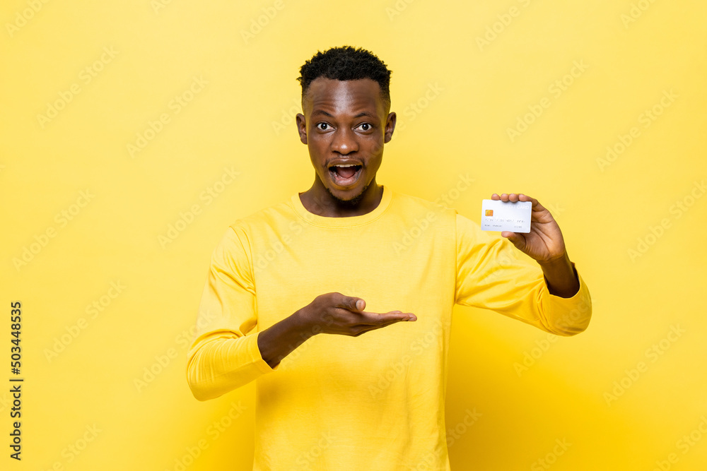Shocked African man in casual attire holding credit card on yellow studio background