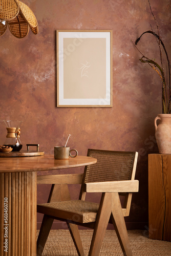 Stylish boho vintage dining room interior with poster mock up. Round table with rattan chair, vase with dried flowers. Brown wall and rattan lamp. Mock up poster. Template. © FollowTheFlow