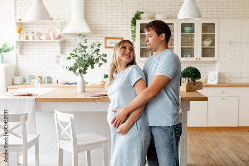 pregnant woman and her handsome husband embracing and smiling while spending time together near window at home. Romantic couple expecting new baby indoors. Parenthood concept