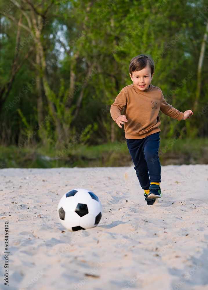 Funny Little Boy Playing with Black and White Soccer Ball in Park on Sand. Kid Running Up to Kick Ball With His Foot