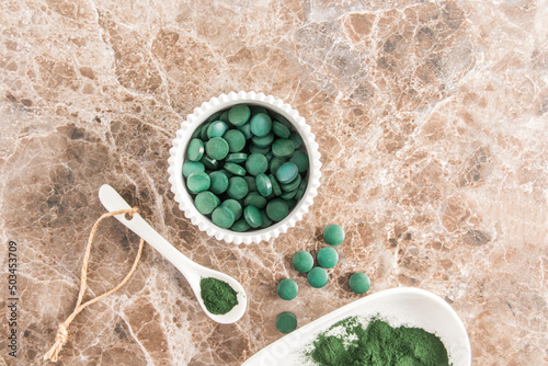 Organic green spirulina powder and green algae tablets in a white bowl and spoon on a marble table. top view. food source of vitamin protein.