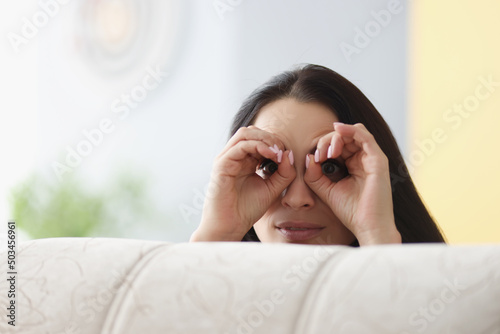 Cheerful woman peeking from behind sofa, female hands make binoculars over eyes photo