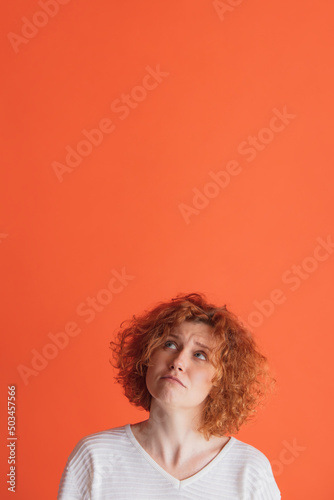 Portrait of emotive red-haired woman posing, looking upwards with houghtful expression isolated over red studio background.