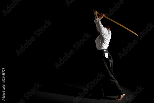 A girl in black hakama standing in fighting pose with wooden sword bokken over black background. Shallow depth of field. 