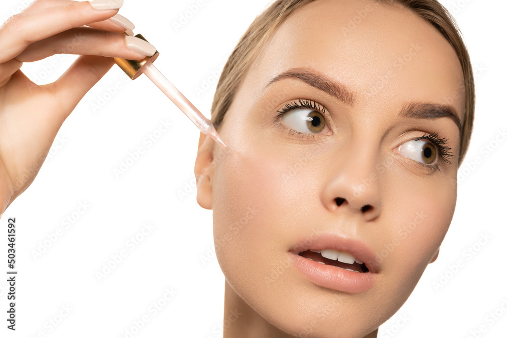 Close up beautiful young woman applying serum, oil on face on white background. Concept of cosmetics, makeup, natural beauty