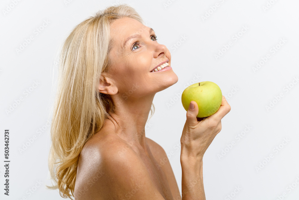 Beautiful and healthy mid age woman with naked shoulders holding green apple near face, looking up and smiling gently. Cosmetology concept Beautiful and healthy mid age woman with naked shoulders holding green apple near face, looking up and smiling gently. Cosmetology concept