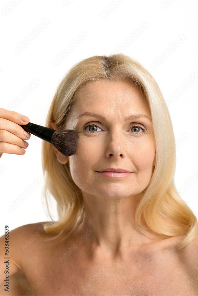 Headshot of mature female is using powder or blush. Serious elegant beautiful middle-aged blonde woman applying make-up with brush isolated on white, caring herself Headshot of mature female is using powder or blush. Serious elegant beautiful middle-aged blonde woman applying make-up with brush isolated on white, caring herself