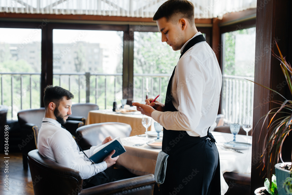 A young businessman in a fine restaurant examines the menu and makes an order to a young waiter in a stylish apron. Customer service. A young businessman in a fine restaurant examines the menu and makes an order to a young waiter in a stylish apron. Customer service.
