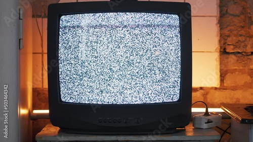 Old flickering CRT TV with antenna and buttons shows interference with scan lines. Refrigerator and locker stand side by side, shabby brick wall with fallen tiles is illuminated by orange neon lamp.