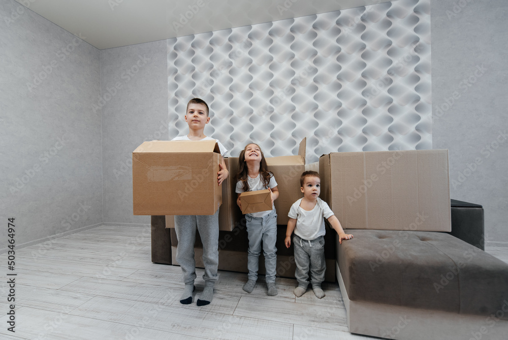 Brothers and sister are playing and enjoying moving to a new apartment against the background of cardboard boxes and a sofa. Purchase of real estate. Housewarming, delivery.