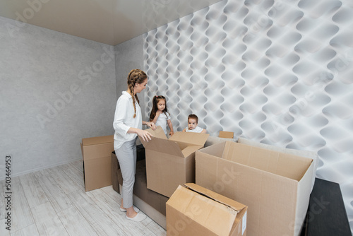 A young mother with children unpacks a box of things after moving to a new apartment. Purchase of real estate. Housewarming, delivery and transportation of goods.