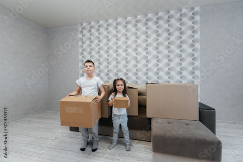 Brothers and sister are playing and enjoying moving to a new apartment against the background of cardboard boxes and a sofa. Purchase of real estate. Housewarming, delivery. © Andrii