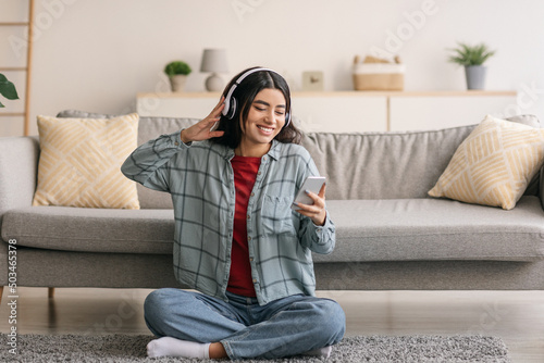 Portrait of cheery young Arab woman in headphones listening to music on mobile device, sitting on floor at home