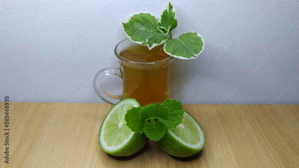 Hand placing mint leaves on top of glass or cup of healthy ginger masala green lime tea with half-sliced lemon on a wooden table with white background and copy space. Horizontal closeup side view.