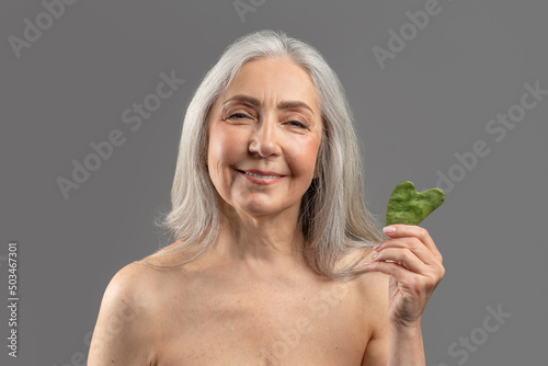 Lovely senior Caucasian woman holding gua sha lymphatic drainage facial self massager and smiling at camera photo