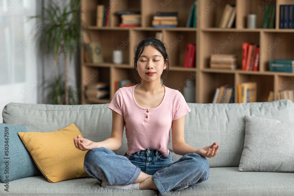 Calm asian lady sitting on comfy sofa and meditating with closed eyes at home, full length, copy space Calm asian lady sitting on comfy sofa and meditating with closed eyes at home, full length, copy space