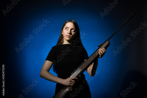 young beautiful woman with long hair in a black t-shirt holding a gun on a blue background