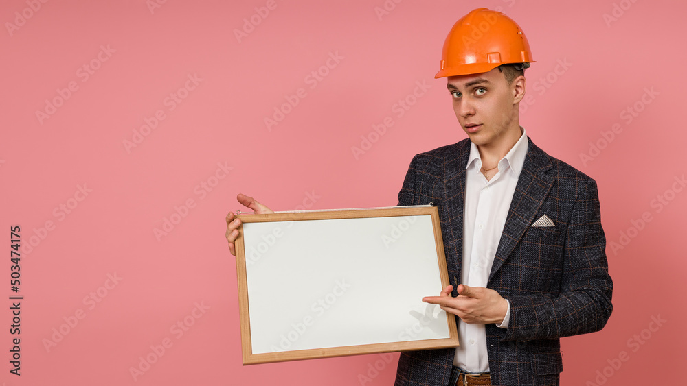 young engineer in a protective helmet and jacket points with a finger at a white board in his hand on a pink background young engineer in a protective helmet and jacket points with a finger at a white board in his hand on a pink background