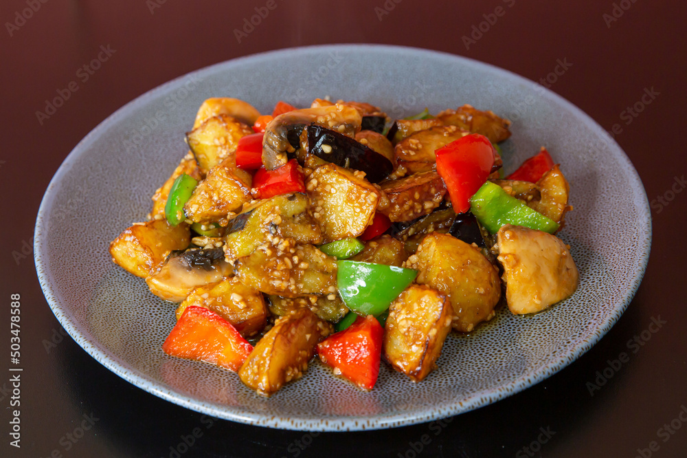 Stir-fried vegetables with eggplant, potato, capsicum of traditional Cantonese yum-cha Asian gourmet cuisine meal food dish on the white serving plate and brown red table Stir-fried vegetables with eggplant, potato, capsicum of traditional Cantonese yum-cha Asian gourmet cuisine meal food dish on the white serving plate and brown red table