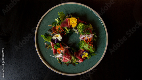 Top view of Tartare beef flower circle salad in the fine dining Japanese restaurant, with a green round plate in black scene reflection table background, raw beef, onion, salad, carrot, flowers