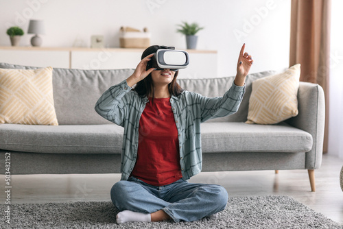 Happy millennial Arab woman in virtual reality glasses exploring cyberspace, playing interactive video game in living room © Prostock-studio
