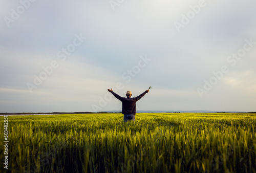 Leinwand Poster Rear view of senior farmer standing in barley field with his outstretched arms at sunset