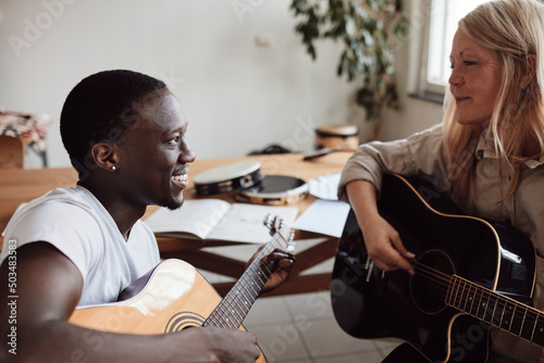 Smiling female mature tutor teaching guitar to young man in classroom