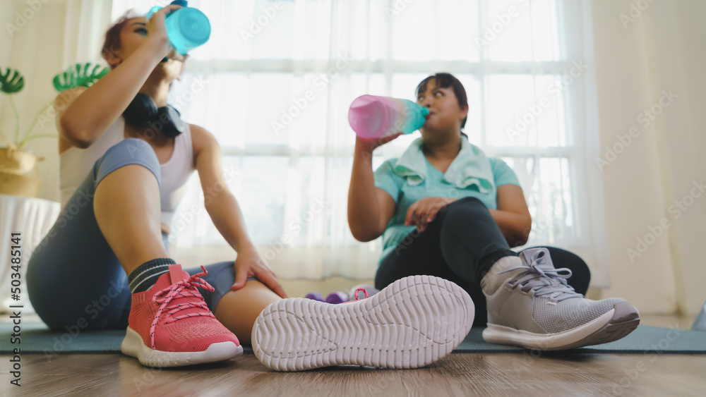 Obraz premium Thirst while exercising concept. Two Asian women's body size is different in sportswear, drinking water while resting and sitting on yoga mat after fitness exercise at home together.