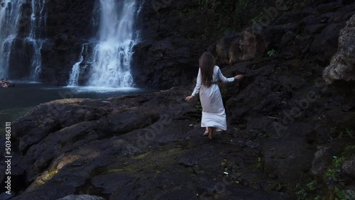 Young beautiful sexy woman in long white dress with long brown hair going on huge stones to waterfall Tad Yuang in Laos. Asia. Tropic forest. Travel Alone with nature. Wind in the Hair 4K Slow motion photo