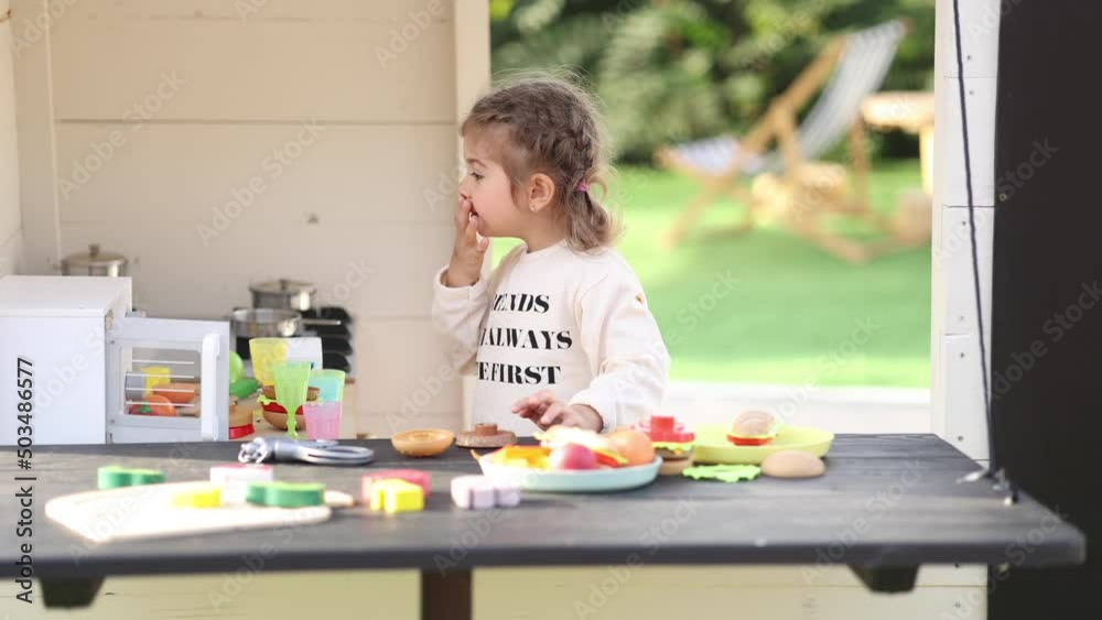 Happy little girl playing on toy kitchen on wheels. Cute girl make a burger on toy kitchen outdoors. Adorable kid play on fresh air Happy little girl playing on toy kitchen on wheels. Cute girl make a burger on toy kitchen outdoors. Adorable kid play on fresh air