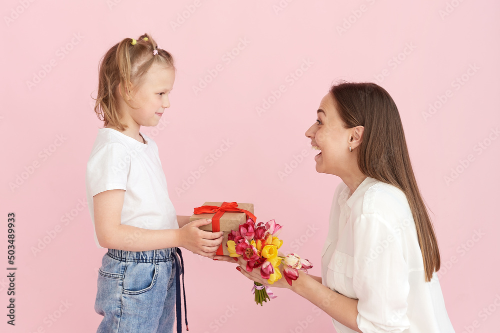 A little daughter gives her mother flowers and a gift for mother's day on a pink background in the studio. Happy mom and daughter portrait A little daughter gives her mother flowers and a gift for mother's day on a pink background in the studio. Happy mom and daughter portrait