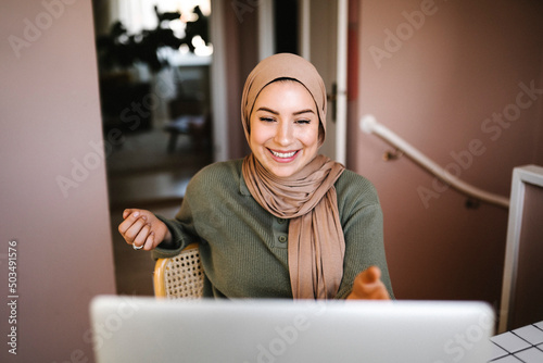 Smiling female freelancer in hijab doing video call through laptop at home photo