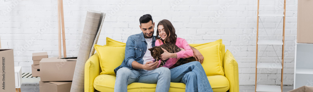 Cheerful interracial couple petting bengal cat on couch near packages at home, banner.