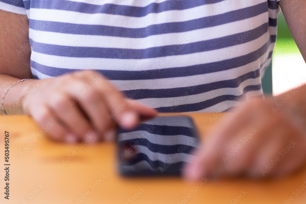 Blurry woman hands using mobile phone resting on the table. Caucasian lady with striped t-shirt sitting at the table Blurry woman hands using mobile phone resting on the table. Caucasian lady with striped t-shirt sitting at the table