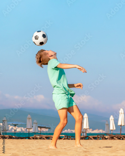 Child boy soccer player performs trick by hitting soccer ball with his head while having fun on sea beach during the summer holidays. Kids games and leisure on sandy seashore on vacation