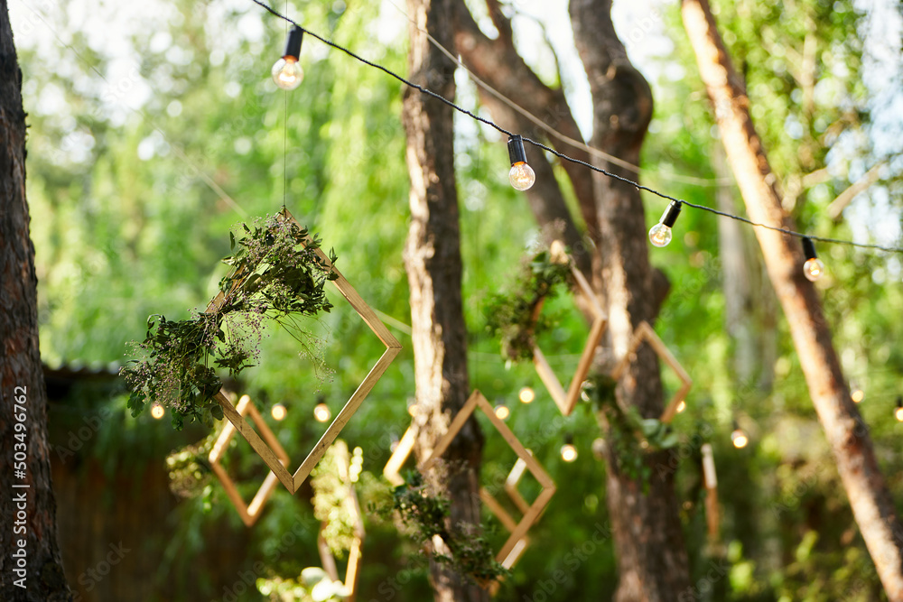 Wedding party banquet outdoors in pine forest. Dining tables, benches decorated in boho style with candles, flowers, spring fairy lights, plates, wineglasses, wooden frames. Eco organic floral decor.