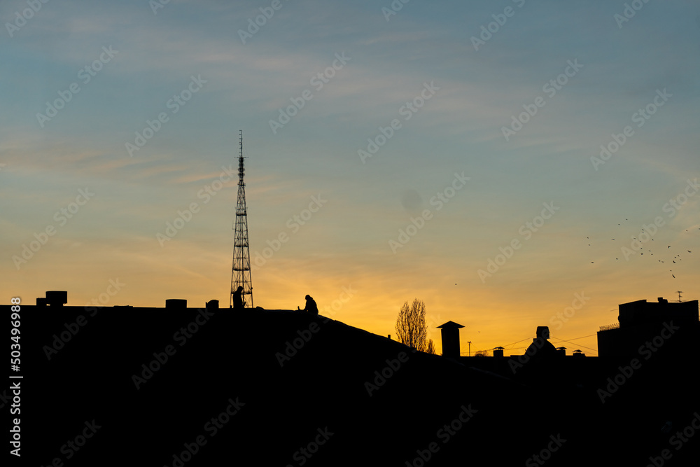 A silhouette of two men working on a roof in the city at a sunset. Golden hour. 
