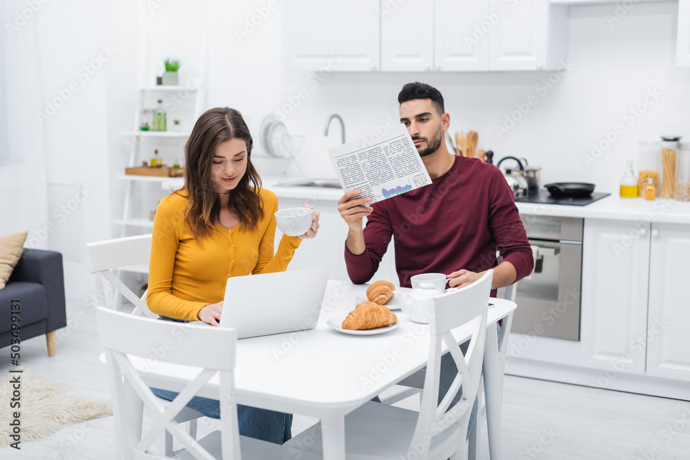 Woman holding cup and using laptop near arabian boyfriend with newspaper in kitchen. Woman holding cup and using laptop near arabian boyfriend with newspaper in kitchen.