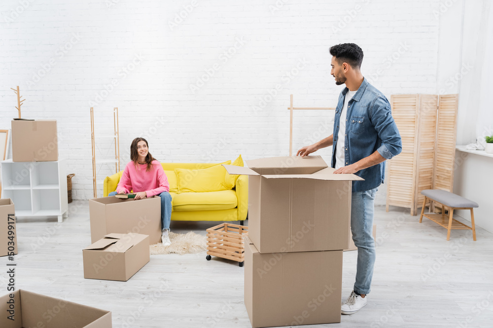 Cheerful woman holding book near arabian boyfriend and packages at home. Cheerful woman holding book near arabian boyfriend and packages at home.
