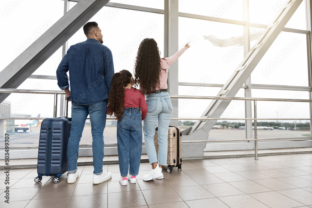 Family Of Three Looking Out Of Window At Airport And Pointing Away Family Of Three Looking Out Of Window At Airport And Pointing Away