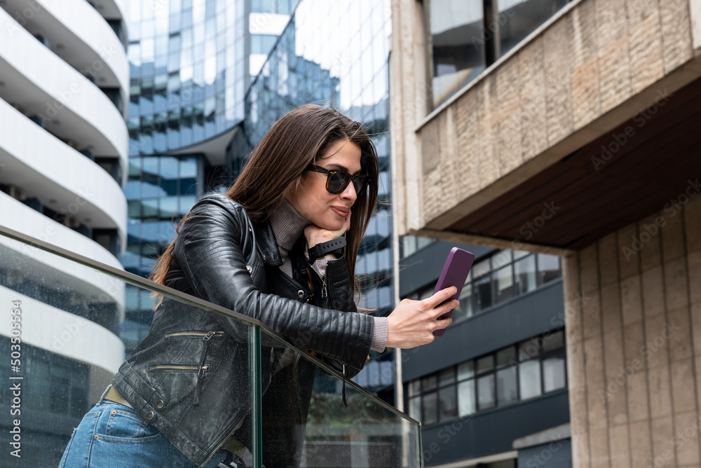 Young beautiful woman in leather jacket and sunglasses waiting for her boyfriend in front of his office building and sending him a text message on his smartphone so they can go to the rock concert.
