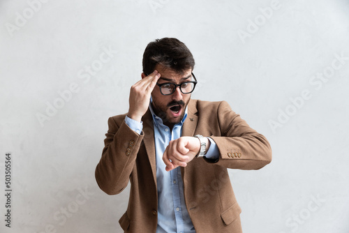 Obraz na płótnie Young business man in suit with eyeglasses with shocked facial expression looking at wristwatch because he is late