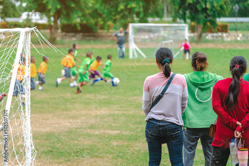 Back view of Moms watch and cheering their sons playing football in school tournament on sideline. Sport, outdoor active,  Spectator watching soccer game. Parents care and encourage their children.