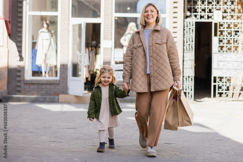 cheerful mother and daughter in trendy clothes walking with shopping bags in city.