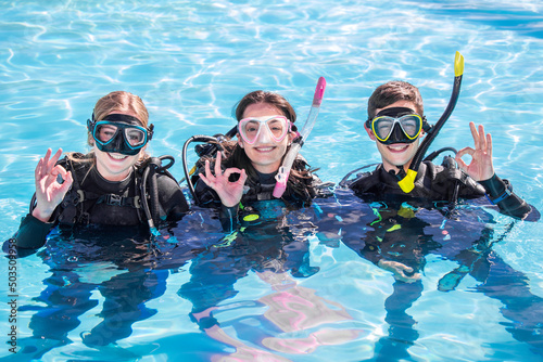 A group of happy scuba divers smiling at the camera with their dive masks and gear on in a pool showing the OK sign