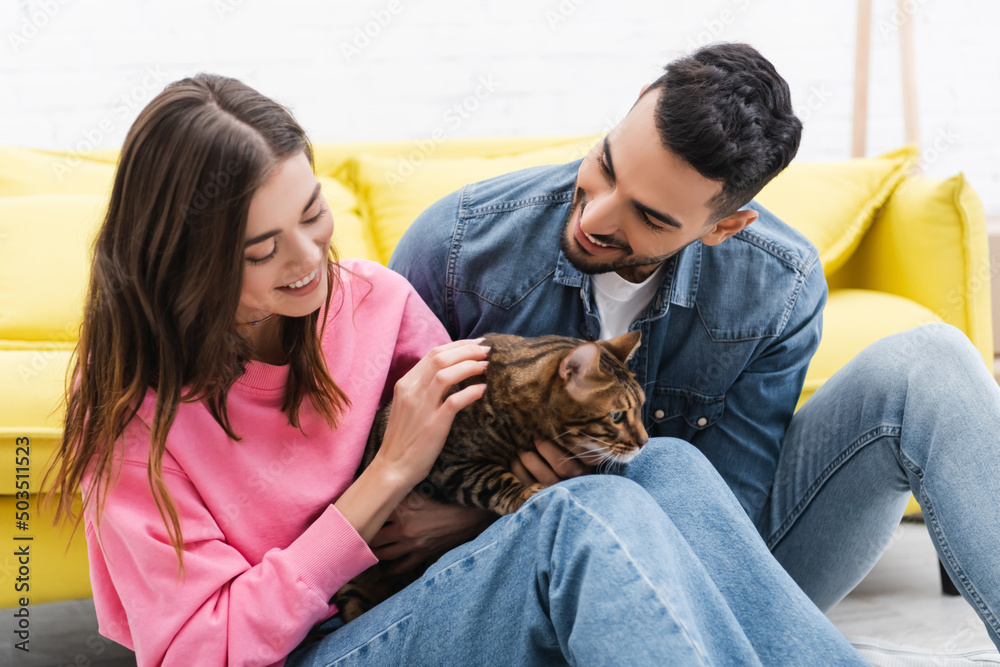 Cheerful multiethnic couple looking at bengal cat at home. Cheerful multiethnic couple looking at bengal cat at home.