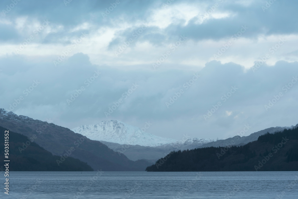 Beautiful Winter landscape image of view along Loch Lomond towards snowcapped mountain range in distance during blue hour