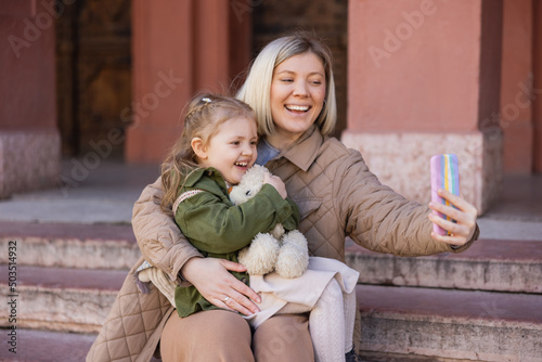 cheerful girl holding teddy bear while mom taking selfie on smartphone.