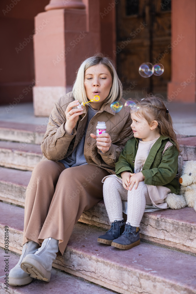 blonde woman blowing soap bubbles while sitting on stairs near daughter.