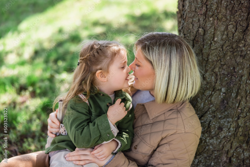 happy mother and child looking at each other while sitting face to face near tree in park.